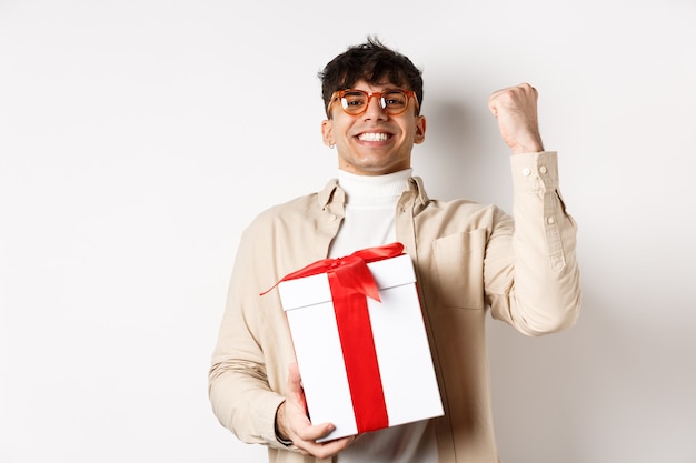 Cheerful guy saying yes as receiving gift, making fist pump and rejoicing, got present, standing on white background.