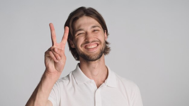 Cheerful guy showing peace sign sincerely smiling at camera over gray background Young positive man posing in studio