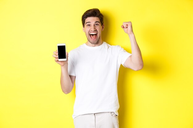 Cheerful guy showing smartphone screen, raising hand up and celebrating, triumphing over internet achievement, standing over yellow background