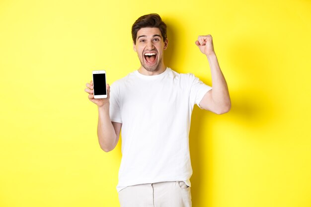 Cheerful guy showing smartphone screen, raising hand up and celebrating, triumphing over internet