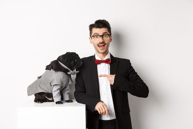 Cheerful guy standing with his cute pug, pointing finger at dog wearing party costume, posing over white background.