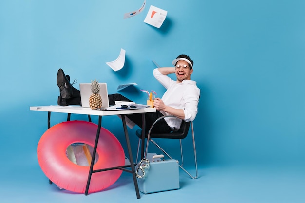 Free photo cheerful guy in white shirt and black pants is working on vacation man in cap enjoys cocktail on blue background with falling documents inflatable circle and suitcase