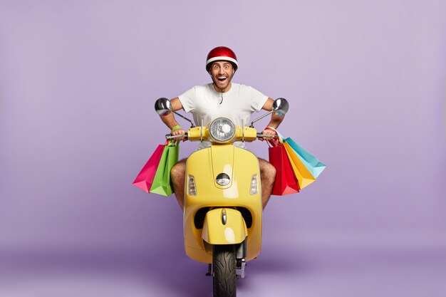 Cheerful guy with helmet and shopping bags driving yellow scooter