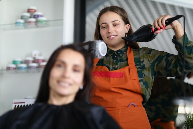 Cheerful hairdresser using hairdryer for treatment with clients hairstyle