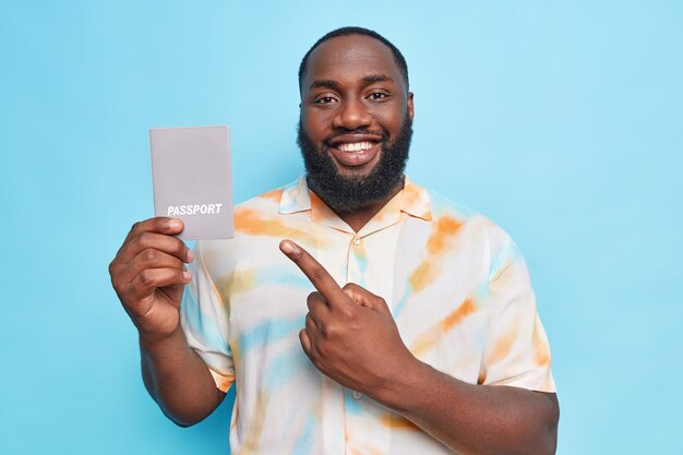 Cheerful handsome bearded man with dark skin points at passport happy about future travel smiles broadly dressed in washed out t shirt isolated over blue wall