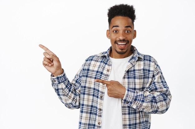 Cheerful handsome Black man, young guy with moustache and stylish hairstyle, pointing fingers left at sale, showing logo banner, smiling happy on white