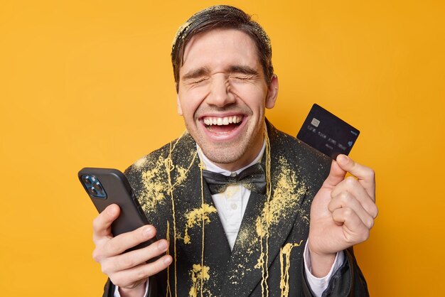 쾌활한 잘생긴 유럽 남자는 행복한 축하를 즐기고 휴대폰을 들고 신용 카드는 온라인 상점에서 구매하는 데 돈을 쓰고 뱀 스프레이로 얼룩진 정장을 입는다