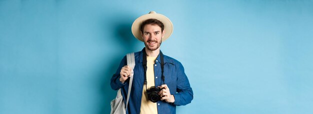 Cheerful handsome guy going on vacation wearing summer hat and holding backpack with camera for phot