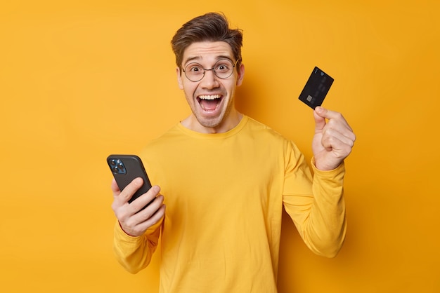 Cheerful handsome guy makes safe mobile banking via cellular application holds credit card provides internet payment wears round spectacles and casual jumper isolated over yellow background