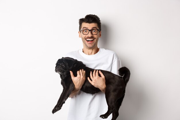 Cheerful handsome man carry black pug and smiling, staring at camera happy. Dog owner holding cute dog in arms, standing over white background.