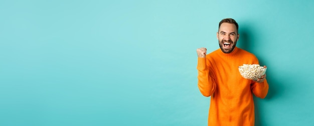 Free photo cheerful handsome man in orange sweater saying yes cheering and rejoicing eating popcorn and watchin