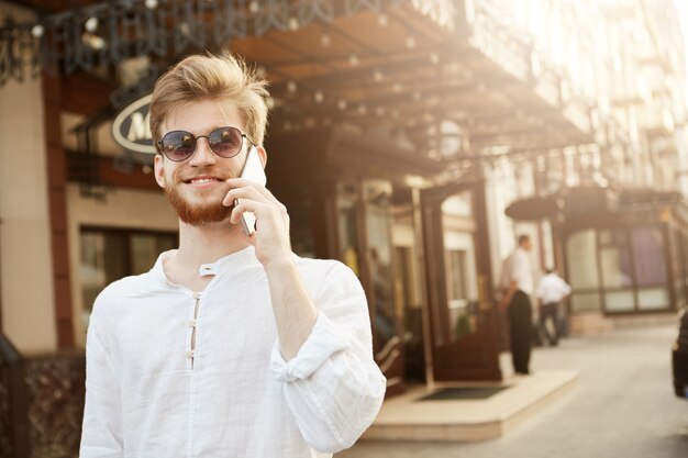 Cheerful handsome red haired guy with trendy hairstyle and beard, in brand new sunglasses talks on the phone with his wife and feeling happy.