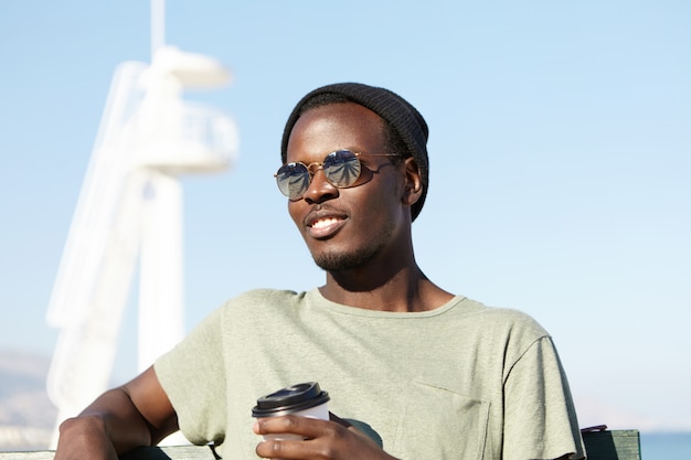 Giovane uomo dalla pelle scura bello allegro che gode della bella giornata estiva in riva al mare, bevendo caffè da asporto, seduto da solo sulla panchina