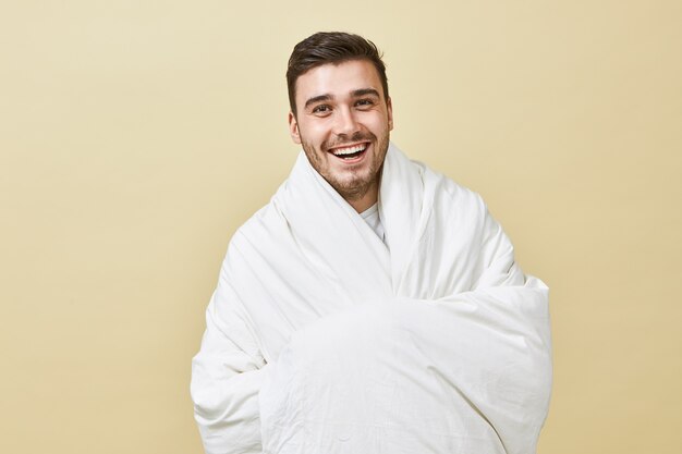 Cheerful happy energetic young male with bristle posing isolated wrapped in white blanket smiling broadly feeling overjoyed and refreshed after good sleep, ready for new day, plans and goals