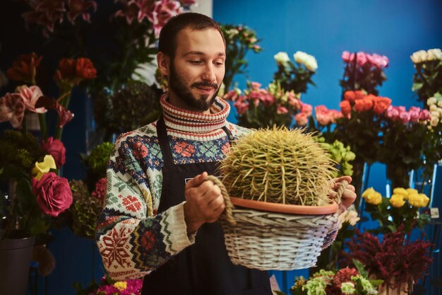 Cheerful happy florist is holding giant cactus in basket at flower shop.