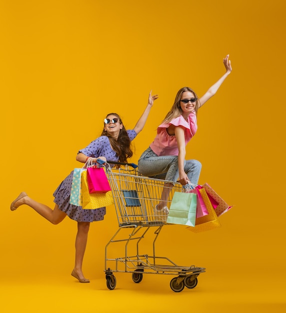 Cheerful happy girls with many shopping bags sitting on shopping cart isolated over yellow studio background