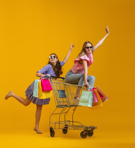 Cheerful happy girls with many shopping bags sitting on shopping cart isolated over yellow studio background