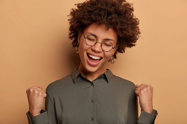 Cheerful happy lucky businesswoman achieves goal, celebrates victory, signing profitable agreement with partner, wears optical glasses and shirt, isolated on brown wall. Student passed test