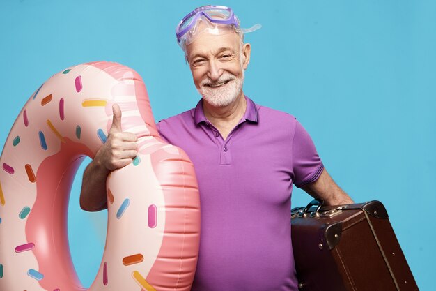 Cheerful happy unshaven male pensioner spending vacations on beach, wearing summer t-shirt and diving scuba mask for snorkeling, holding suitcase and inflatable swimming ring, smiling excitedly