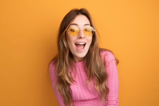 Cheerful and happy young beautiful woman in pink top wearing glasses looking at camera smiling standing over orange background