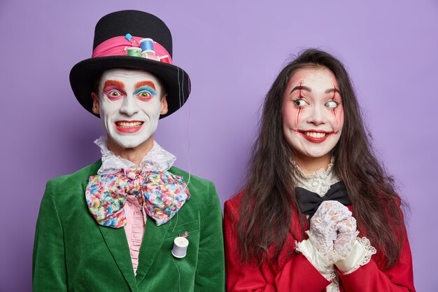 Cheerful hatter in green costume bowtie and tall hat cheerful spooky woman wears scary makeup isolated on purple wall