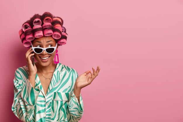 Cheerful hesitant housewife talks on phone, listens rumors and gets pleasant offer wears hair curlers, sunglasses and dressing gown, looks aside happily poses over rosy wall blank space for text