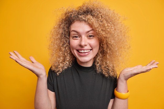 Cheerful hesitant woman with curly hair spreads palms feels reluctant and uncertain smiles joyfully wears casual black t shirt isolated over yellow wall. Doubtful unsure glad female model