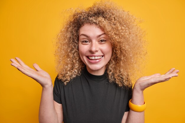 Cheerful hesitant woman with curly hair spreads palms feels reluctant and uncertain smiles joyfully wears casual black t shirt isolated over yellow wall. Doubtful unsure glad female model