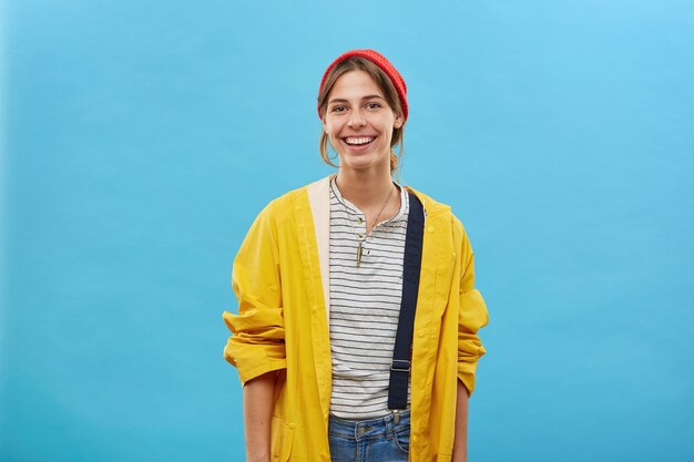 Cheerful hipster girl in stylish yellow raincoat and red hat