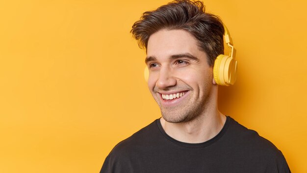 Cheerful hipster guy with dark hair smiles positively listens favorite audio track looks away poses against yellow background copy space for your promotional content Stereo accessory and technology