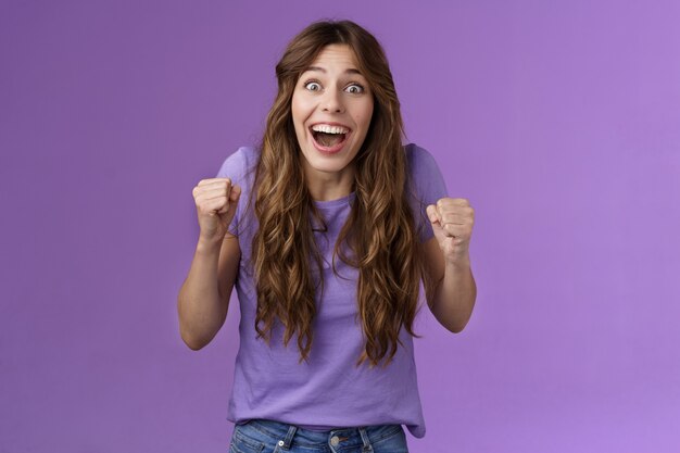 Cheerful hopeful cute female fan celebrating awesome news winning lottery yelling pleased happy smiling broadly fist pump joyfully happiness success triumph pose purple background