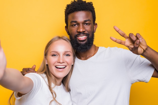 Cheerful interrational couple taking self-portrait together, looking at front and smiling, posing over yellow wall