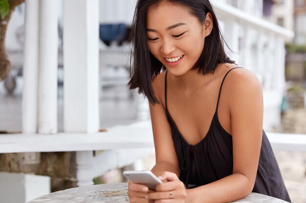 Cheerful Japanese woman installs new application on mobile phone while waits for dish in restaurant, connected to wireless internet, has happy expression. People, leisure and technology concept