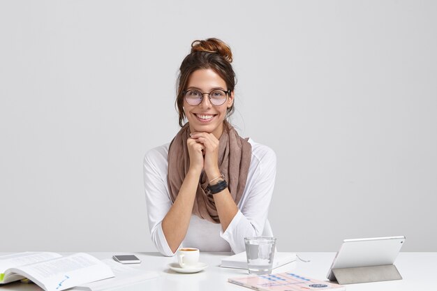 Cheerful journalist with pleasant appearance, wears transparent glasses, has dark haired combed, enriches her knowledge while reading, has tender smile sits at workspace with touchpad, glass of water