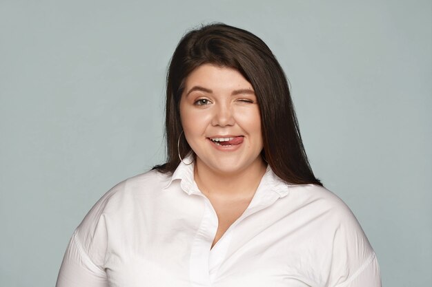 cheerful joyful chubby overweight female in formal white shirt having fun at office during break, winking and sticking out tongue, flirting or teasing you