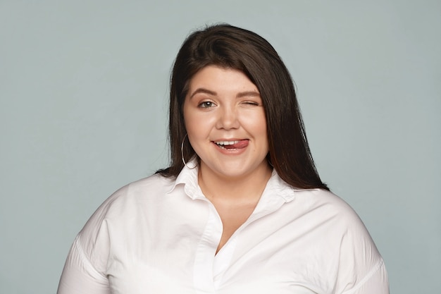 Free photo cheerful joyful chubby overweight female in formal white shirt having fun at office during break, winking and sticking out tongue, flirting or teasing you