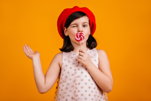 Веселый ребенок ест леденец на палочке. короткошерстная девочка десятилетнего возраста с конфетой, изолированной на желтом.