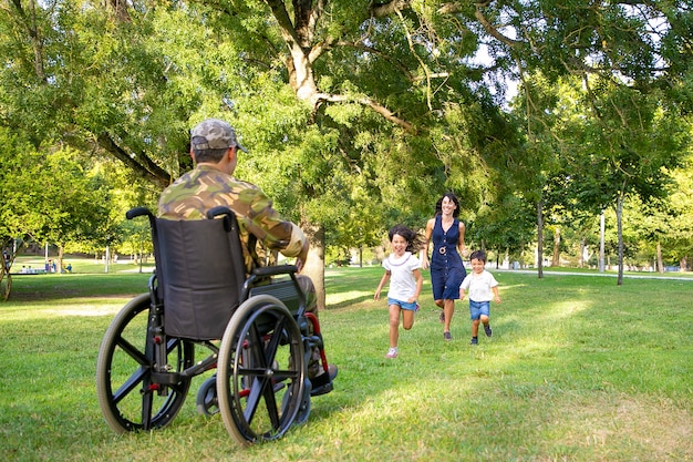 Cheerful kids and their mom meeting military father and running to disabled man in camouflage. Veteran of war or returning home concept