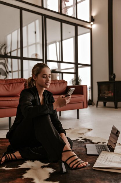 Cheerful lady in black silk home suit sits on carpet smiles and reads message in cellphone Attrac