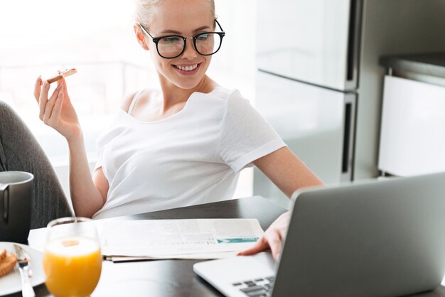 Cheerful lady eating bread with jam and using laptop