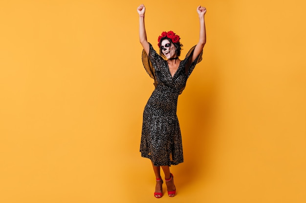 Cheerful lady emotionally dances with her arms raised up. Portrait of model with dark hair and crown of roses in Halloween outfit.