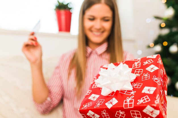 Cheerful lady holding gift box and credit card near Christmas tree