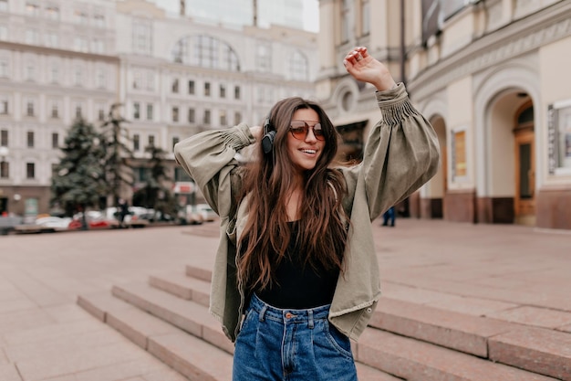 Cheerful lady is listening music and dancing in the city Pretty young girl with long dark hair in jacket and jeans with nude makeup smiling and narrowing eyes while waving head