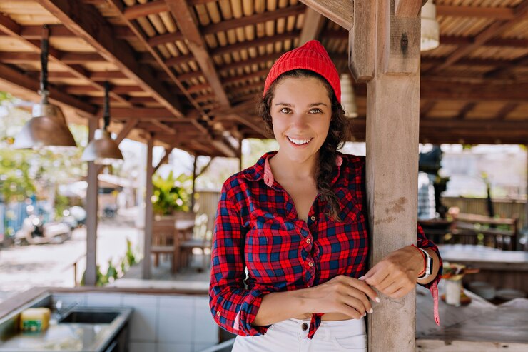 Cheerful lady posing at camera on background of wood open space cafe. Female tourist has fun on sunny summmer day. Concept of single trip holidays and happiness