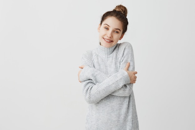 Free photo cheerful lady standing with positive facial expressions. good-looking female interior designer hugging herself feeling warm in woolen grey dress. sensation concept