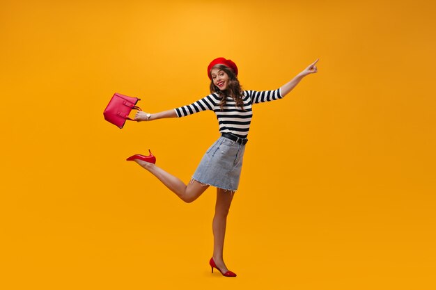 縞模様のシャツ、赤いベレー帽jumping.nオレンジ色の背景の陽気な女性。 i番目のハンドバッグ。ベレー帽とトレンディな靴のポーズで魅力的な若い女性。