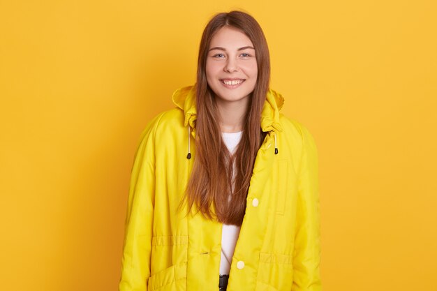 Cheerful lady wearing bright yellow jacket , woman has long beautiful hair, being in good mood, standing with happy facial expression.