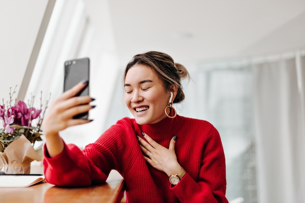 Cheerful lady in wireless earphones wearing red sweater talking on phone and laughing while sitting by window