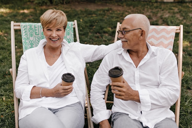 Cheerful lady with blonde short hair in white blouse laughing, holding cup of tea and posing with old man in eyeglasses outdoor.