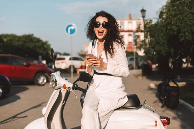 Signora allegra con capelli ricci scuri in costume bianco e occhiali da sole seduta su un motorino bianco con il cellulare in mano e felicemente guardando a porte chiuse sulla strada con vista sulla città sullo sfondo